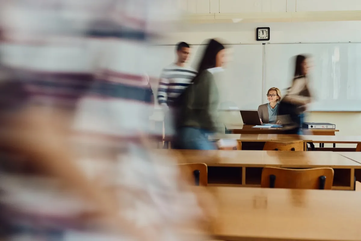 Teacher seated at desk with laptop smiling as motion-blurred students walk through a classroom